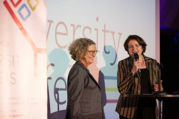 Two women on a stage, one speaking into a microphone, a colorful logo and the word "Diversity" in the background.