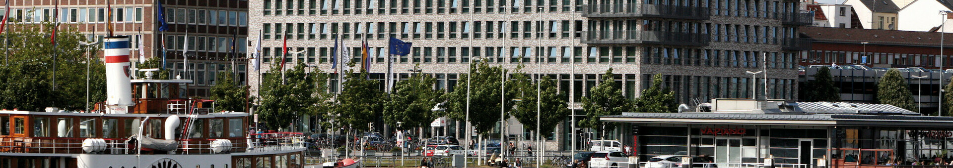 Außenansicht des ATLANTIC Hotel Kiel Moderne Hotelgebäude am Wasser mit einem historischen Dampfschiff im Vordergrund, blauer Himmel im Hintergrund.