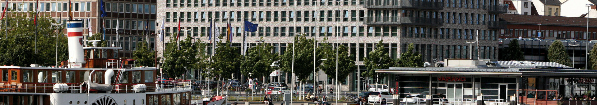 Exterior view of the ATLANTIC Hotel Kiel Modern hotel building on the waterfront with a historic steamship in the foreground, blue sky in the background.