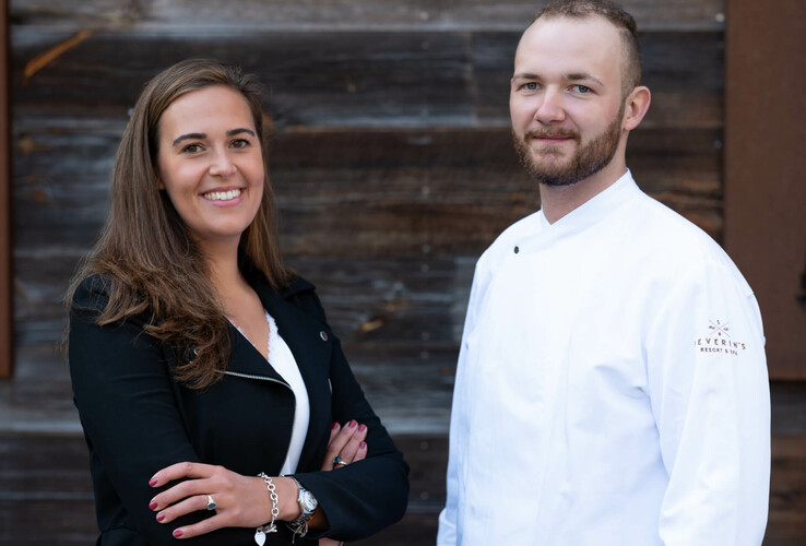 Zwei lächelnde Personen, eine Frau in schwarzem Blazer und ein Mann in weißer Kochjacke, vor Holzwand.