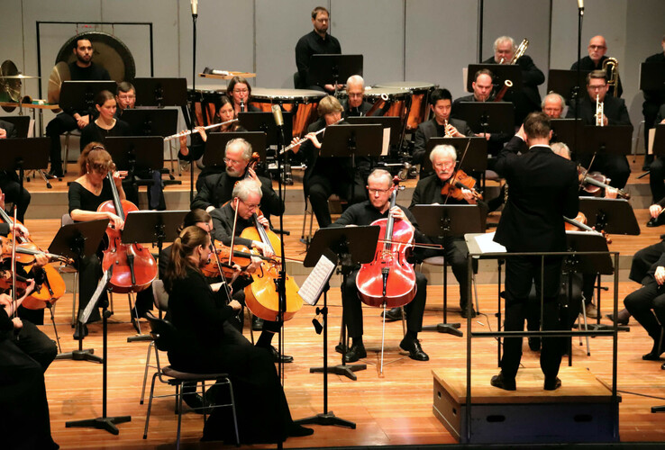 Ein Orchester spielt auf einer Bühne, dirigiert von einem Dirigenten. Musiker mit Streich- und Blasinstrumenten.