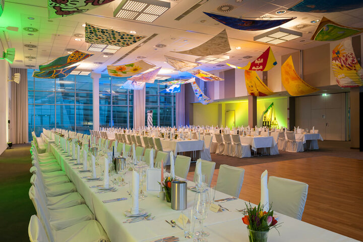 Eleganter Bankettsaal im ATLANTIC Hotel Sail City, Bremerhaven, mit langen, festlich gedeckten Tischen und bunter Deckendekoration.