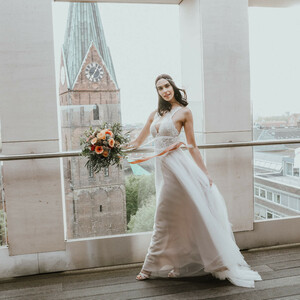 Braut in weißem Kleid mit Blumenstrauß, steht vor Panoramafenster mit Kirchturm und Stadtblick im Hintergrund.