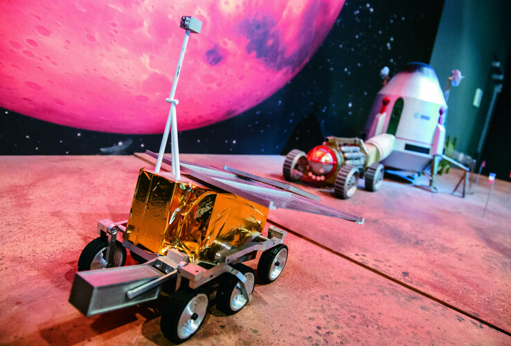 Model of a lunar rover and a space capsule in front of a large, pink-colored planetary background.