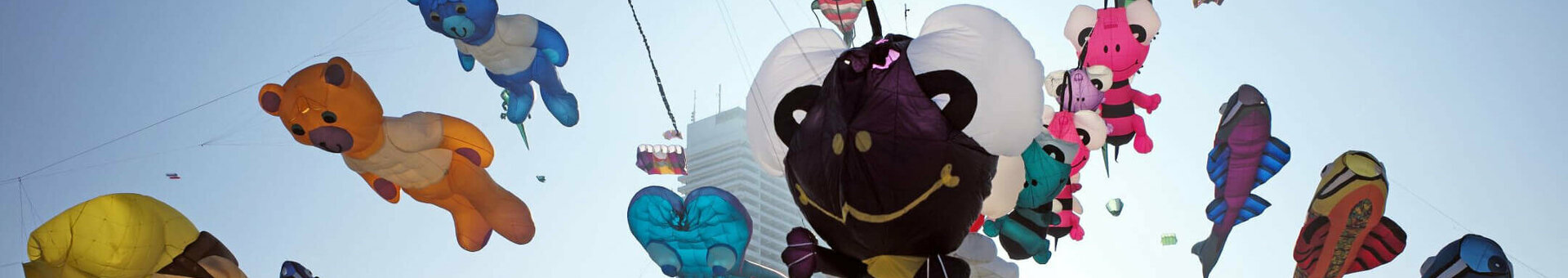 Bunte Drachen in Tier- und Figurenform fliegen über einem Strand, im Hintergrund ein hohes Gebäude.