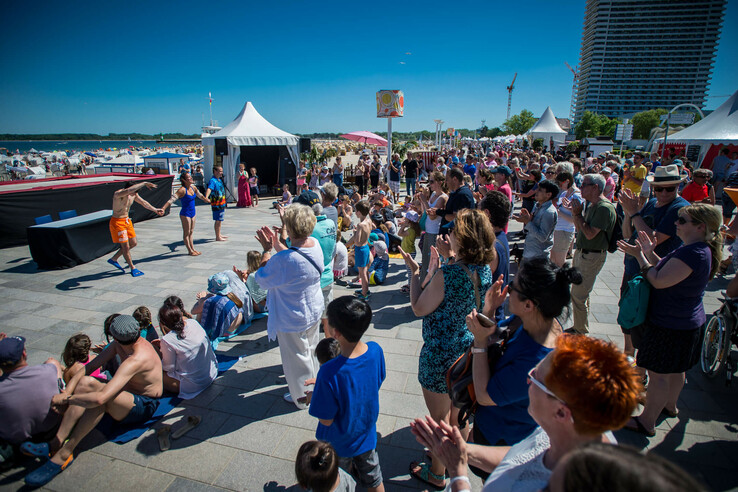 Menschenmenge applaudiert einer Strandaufführung bei Sonnenschein, im Hintergrund das Meer und ein Hotelgebäude.