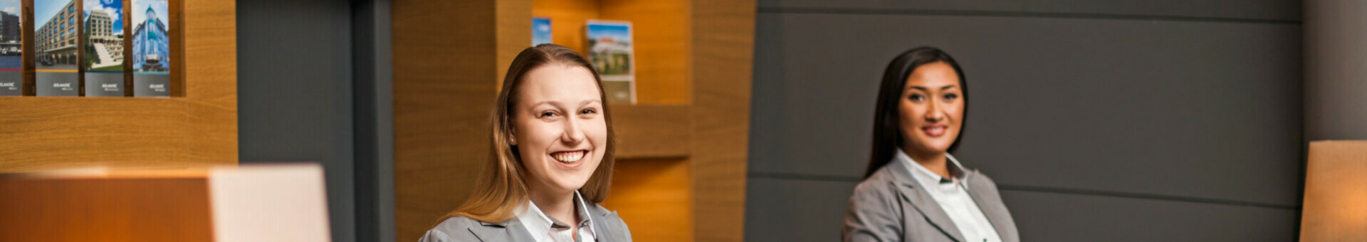 Rezeption im ATLANTIC Hotel Airport in Bremen  Zwei Empfangsmitarbeiterinnen in grauen Anzügen lächeln an der Rezeption des ATLANTIC Hotel Airport Bremen.