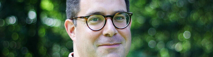 Hoteldirektor Sascha von Zabern Mann mit Brille und rosa Polo-Shirt vor unscharfem, grünem Hintergrund im Freien.