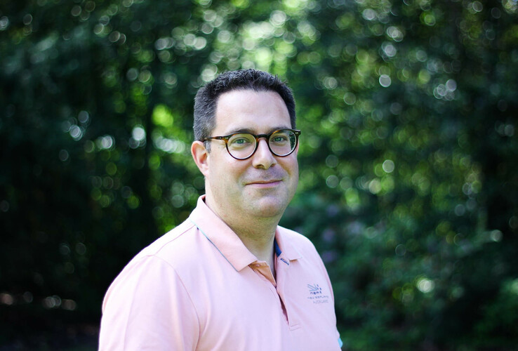 Mann mit Brille und rosa Polo-Shirt vor unscharfem, grünem Hintergrund im Freien.