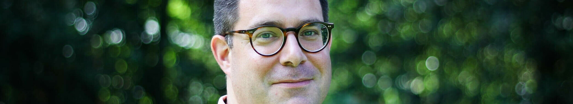 Mann mit Brille und rosa Polo-Shirt vor unscharfem, grünem Hintergrund im Freien.