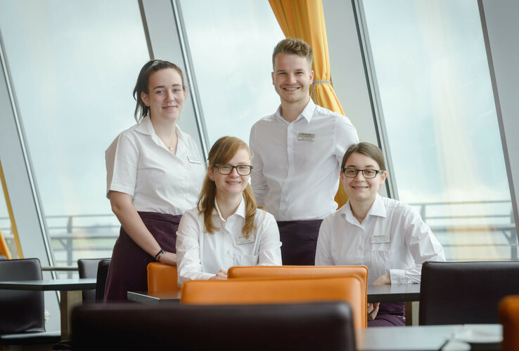 Hotel staff in white clothing smiling in a modern dining room with large windows and orange chairs.
