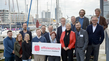 Gruppe von Personen posiert mit einem Schild am Hafen, Boote und moderne Gebäude im Hintergrund.