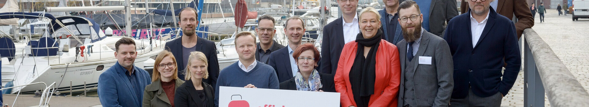 Gruppe von Personen posiert mit einem Schild am Hafen, Boote und moderne Gebäude im Hintergrund.