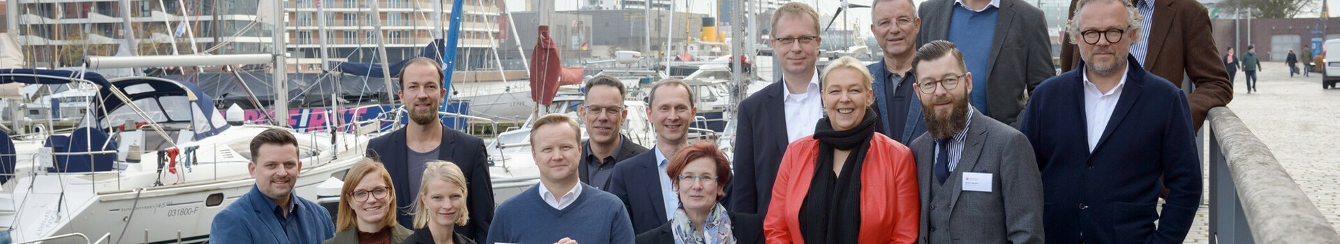 Energiekonsens Hotellerie Gruppe von Personen posiert mit einem Schild am Hafen, Boote und moderne Gebäude im Hintergrund.