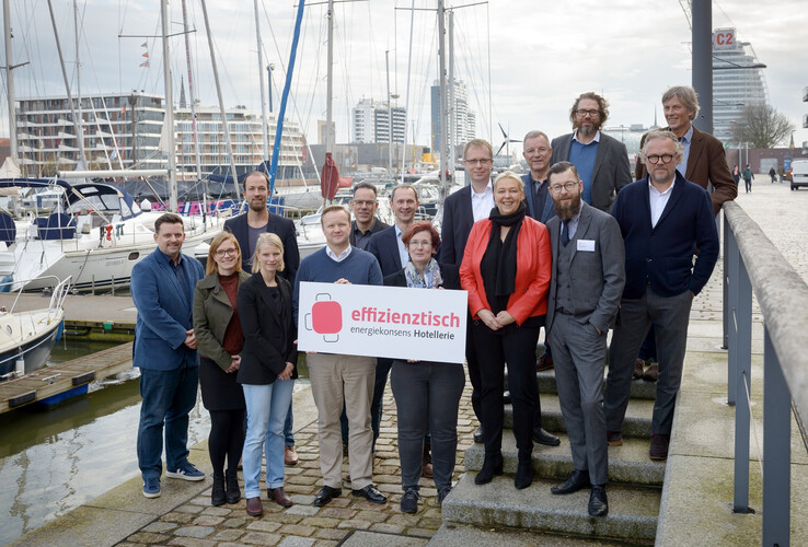 Gruppe von Personen posiert mit einem Schild am Hafen, Boote und moderne Gebäude im Hintergrund.