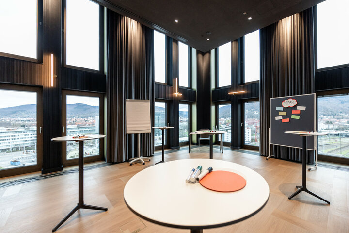 Moderner Konferenzraum im ATLANTIC Hotel Heidelberg mit großen Fenstern, Stehtischen, Flipchart und Blick auf die Stadt.