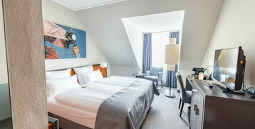Helles Hotelzimmer mit Doppelbett, Schreibtisch, TV, Sessel und großem Fenster im ATLANTIC Hotel Vegesack, Bremen.