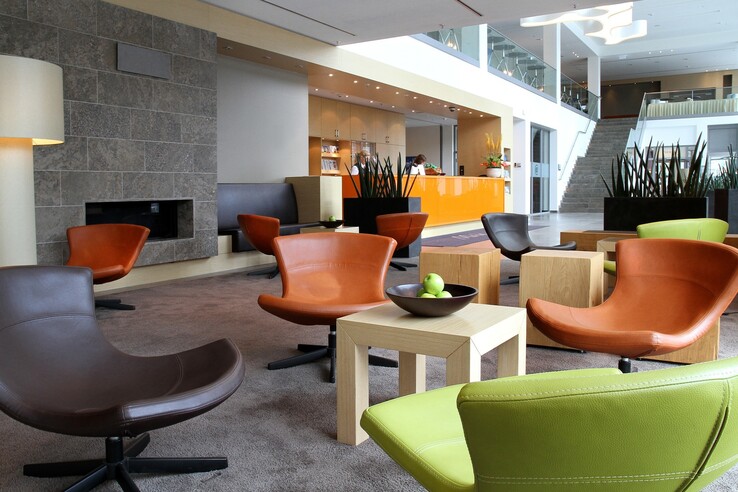 Moderne Hotel-Lobby mit bunten Sesseln, Holztischen und einem orangefarbenen Empfangstresen im ATLANTIC Hotel Kiel.
