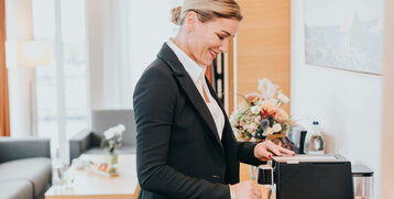 Frau bereitet Kaffee in modernem Hotelzimmer des ATLANTIC Hotel Kiel zu, im Hintergrund gemütliche Einrichtung.