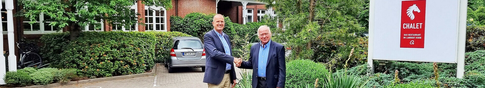 Zwei Männer schütteln Hände vor dem Atlantic Hotel Landgut Horn, umgeben von grüner Landschaft.