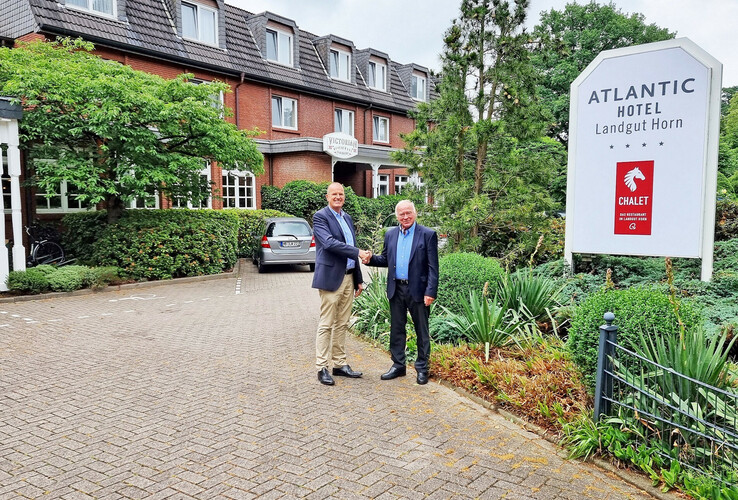 Zwei Männer schütteln Hände vor dem Atlantic Hotel Landgut Horn, umgeben von grüner Landschaft.