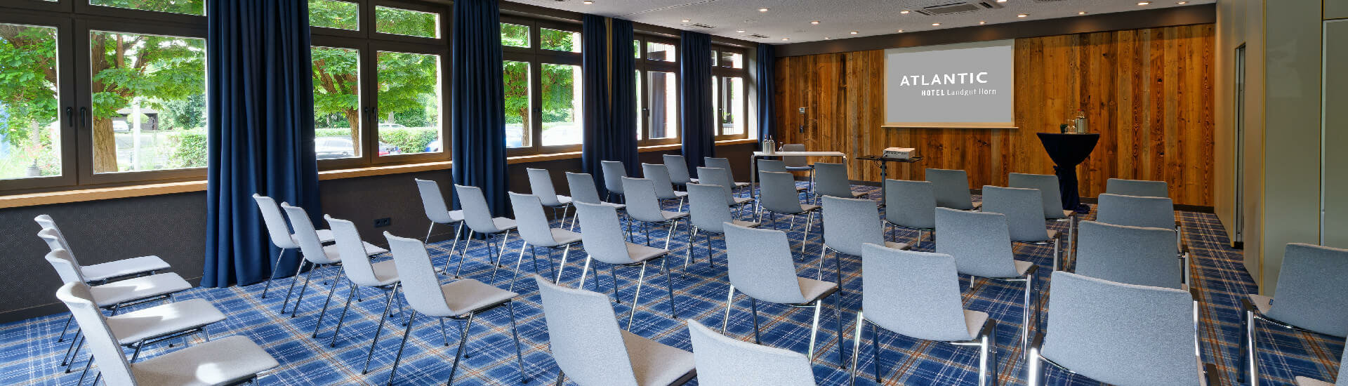 Moderner Konferenzraum im ATLANTIC Hotel Landgut Horn mit blauen Stühlen, Holzwand und großen Fenstern.
