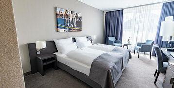 Helles Hotelzimmer mit Doppelbett, Sitzbereich, Schreibtisch und großem Fenster mit Vorhängen im ATLANTIC Hotel Vegesack.