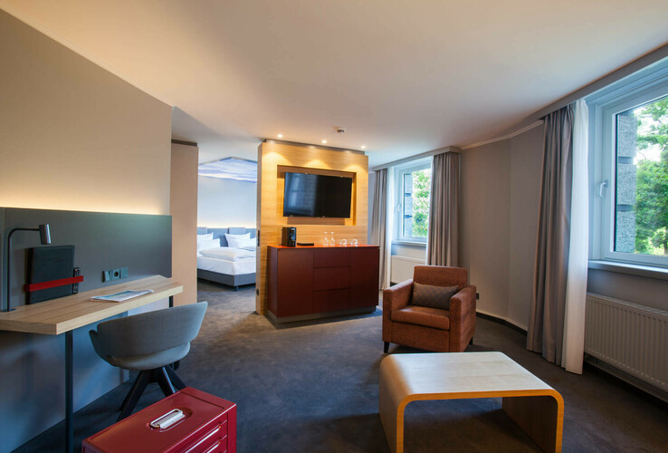 Modernes Hotelzimmer mit Sitzbereich, Schreibtisch, TV, großem Fenster und Blick auf das Schlafzimmer im ATLANTIC Hotel Airport.