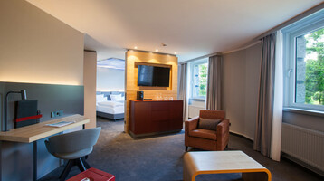 Modern hotel room with seating area, desk, TV, large window and view of the bedroom at the ATLANTIC Hotel Airport.