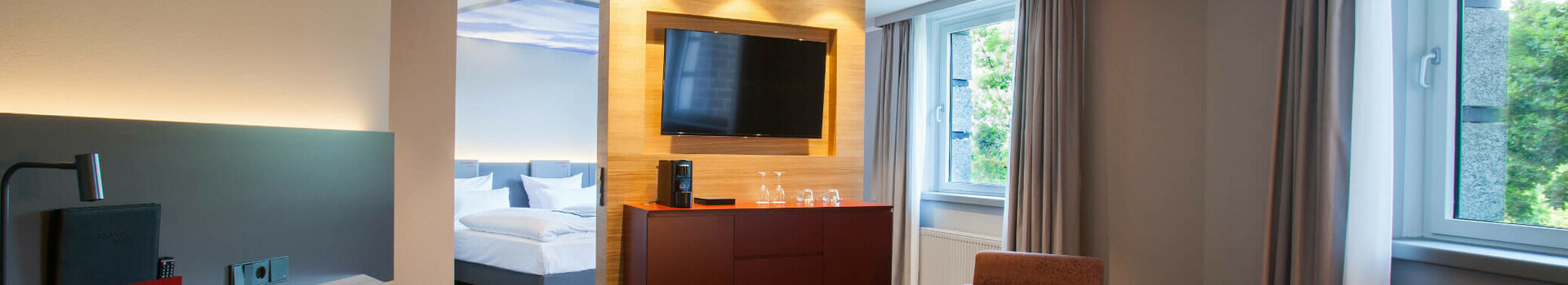 ATLANTIC Hotel Airport Bremen Suite Piccard Modernes Hotelzimmer mit Sitzbereich, Schreibtisch, TV, großem Fenster und Blick auf das Schlafzimmer im ATLANTIC Hotel Airport.