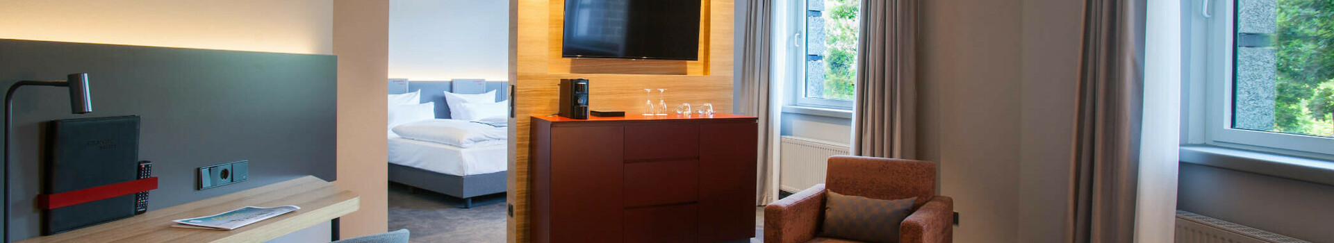 ATLANTIC Hotel Airport Bremen Suite Piccard Modernes Hotelzimmer mit Sitzbereich, Schreibtisch, TV, großem Fenster und Blick auf das Schlafzimmer im ATLANTIC Hotel Airport.