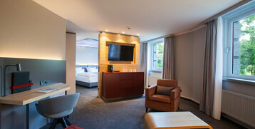 Modernes Hotelzimmer mit Sitzbereich, Schreibtisch, TV, großem Fenster und Blick auf das Schlafzimmer im ATLANTIC Hotel Airport.