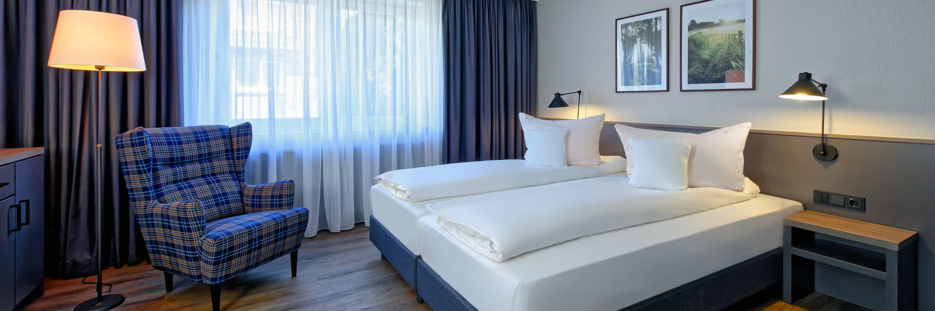 Bright hotel room with double bed, comfortable armchair, floor lamp and two pictures on the wall.