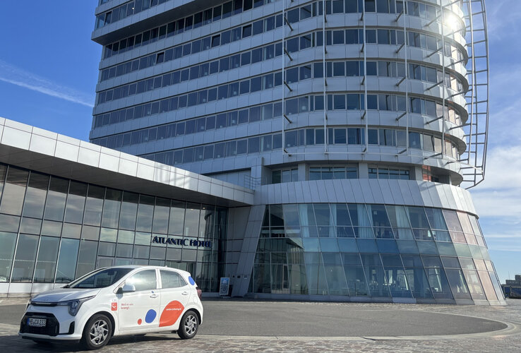 Ein weißer Kia Rio mit cambio-Logo steht vor dem ATLANTIC Hotel Sail City