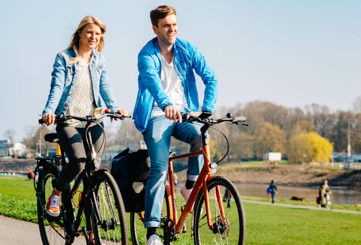Paar fährt bei sonnigem Wetter auf einem Radweg am Fluss entlang, umgeben von grünem Gras und Bäumen.