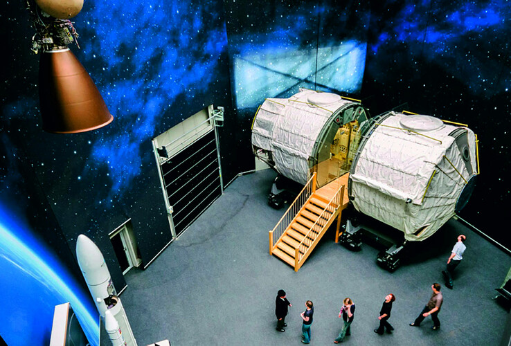 Interior view of a space module with stairs, surrounded by space decorations, people looking at the exhibition.