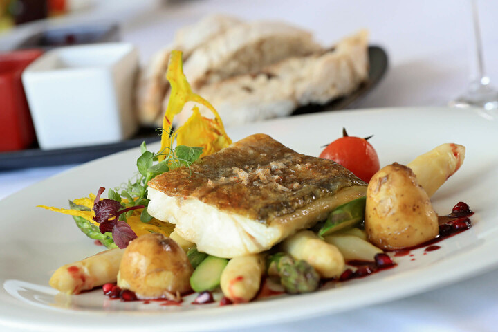 Speisen und Getränke im ATLANTIC Grand Hotel Travemünde Gegrillter Fisch auf Spargel und Kartoffeln, garniert mit Kirschtomate und Kräutern, auf weißem Teller serviert.