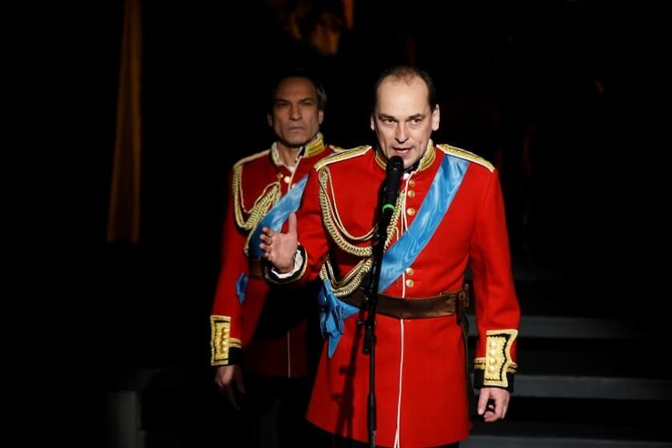 Two men in red uniforms with blue sashes speak into microphones on a stage. © bremer shakespeare company