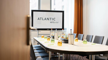 Moderner Konferenzraum im ATLANTIC Hotel Kiel mit gedecktem Tisch und Präsentationsbildschirm.