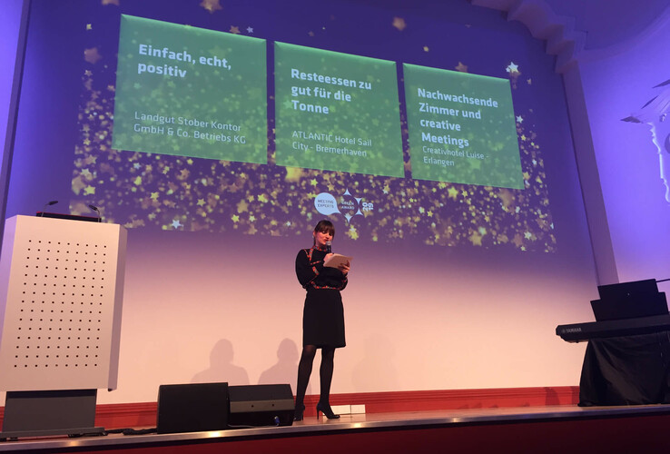 A woman gives a presentation on a stage with projected texts about the ATLANTIC Hotel Sail City in Bremerhaven.