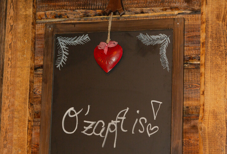 Tafel mit Schriftzug "O'zapft is!" und rotem Herzanhänger, rustikaler Holzrahmen.