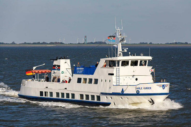 Weiße Fähre auf offenem Meer, deutsche Flagge, mehrere Passagiere an Deck, im Hintergrund Küste mit Windrädern.