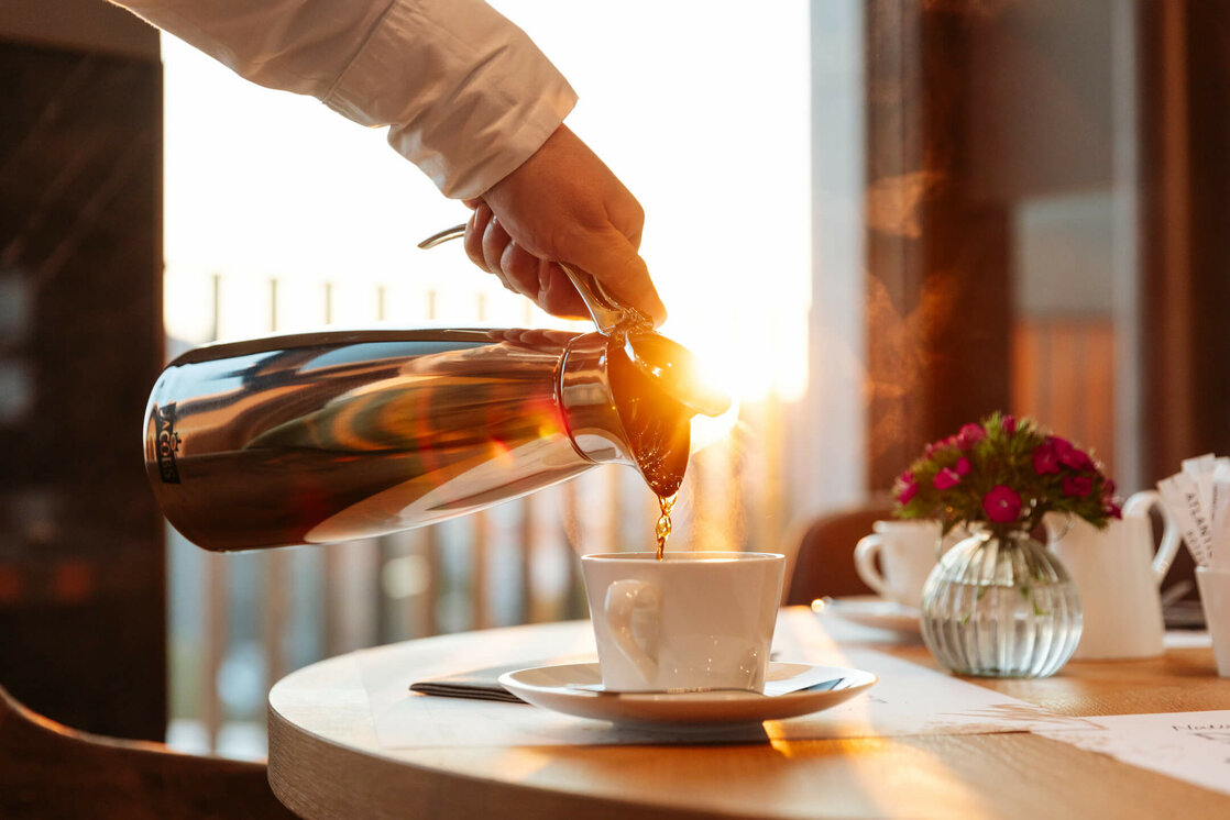 ATLANTIC Hotel Heidelberg Breakfast Coffee Coffee is poured into a cup on a table with flowers and sunlight in the background.
