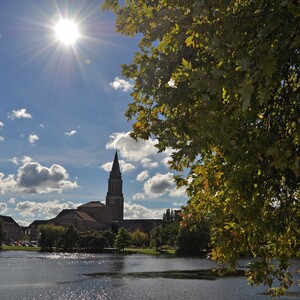 Sonniger Tag am See mit Kirchturm und grünen Bäumen im Vordergrund, Wolken am blauen Himmel.