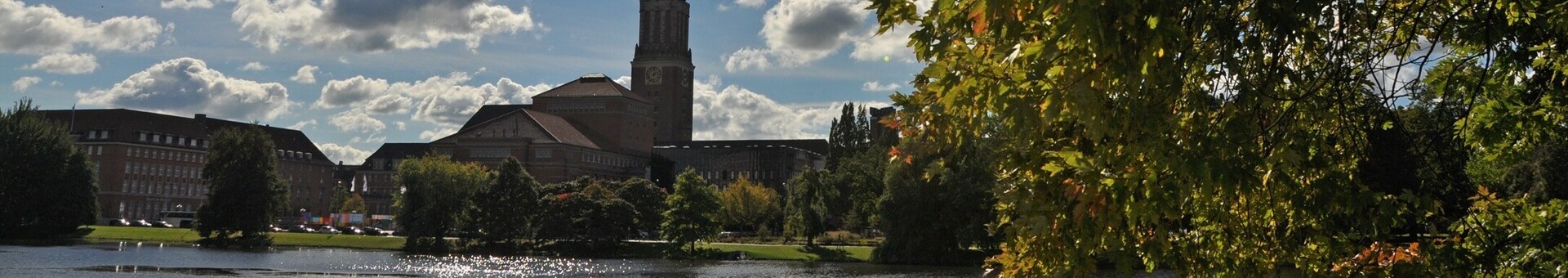 Sonniger Tag am See mit Kirchturm und grünen Bäumen im Vordergrund, Wolken am blauen Himmel.
