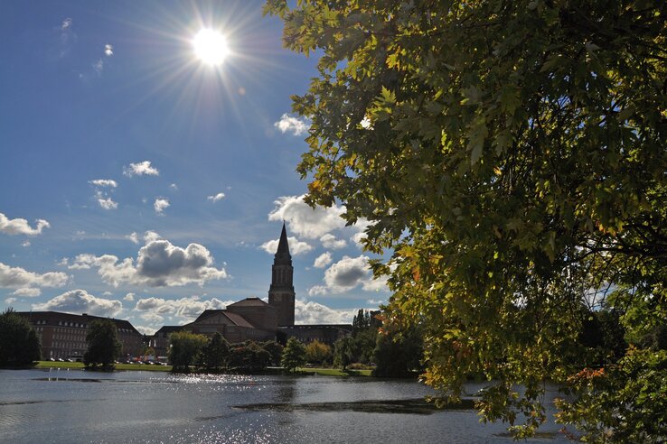 Sonniger Tag am See mit Kirchturm und grünen Bäumen im Vordergrund, Wolken am blauen Himmel.