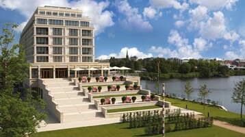 Modern hotel by the river with terraces, sun loungers and parasols, surrounded by green countryside and blue skies.