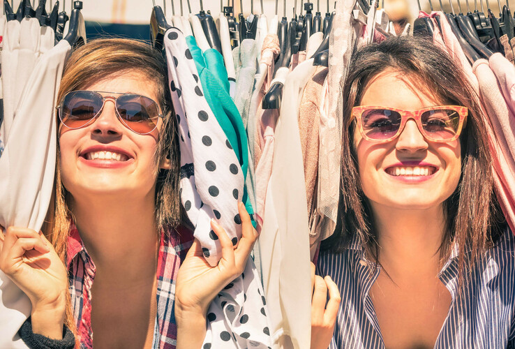 Zwei lachende Frauen mit Sonnenbrillen zwischen bunten Kleidungsstücken auf einem Kleiderständer.