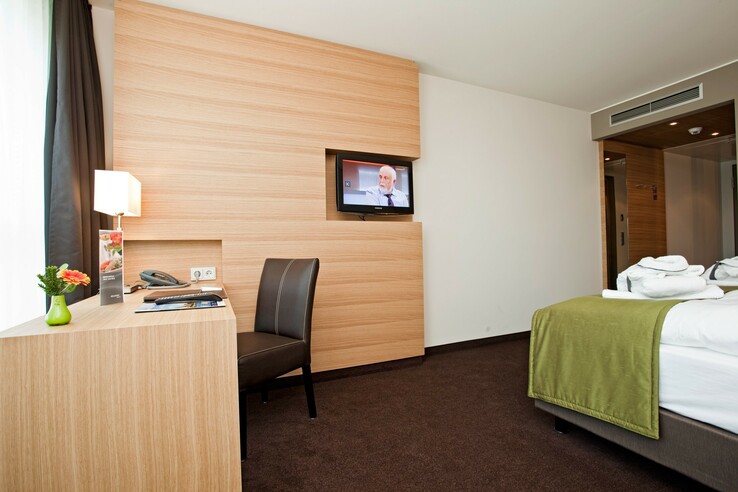 Modernes Hotelzimmer mit Schreibtisch, Stuhl, Flachbild-TV und Bett mit grüner Decke im ATLANTIC Hotel Kiel.