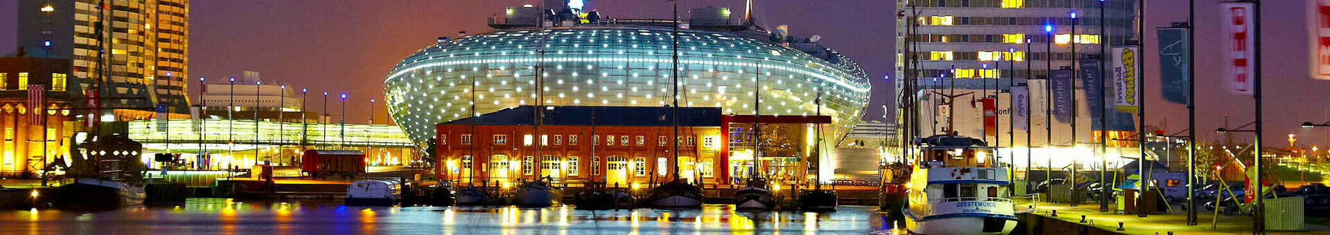 Bremerhaven at night with illuminated ATLANTIC Hotel Sail City on the waterfront, surrounded by buildings and boats.